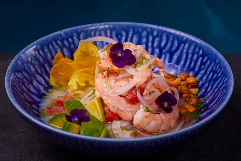 Ceviche de gambas servido en un bol azul, acompañado de aguacate, chips de plátano y maíz tostado, en El Ñaño Internacional
