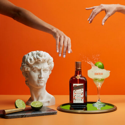 Fotografía creativa de producto: botella de Cointreau sobre bandeja dorada con un cóctel Margarita, acompañada de elementos artísticos como un busto de mármol y limas frescas, todo en un fondo naranja vibrante. Imagen realizada por [nombre de tu estudio fotográfico